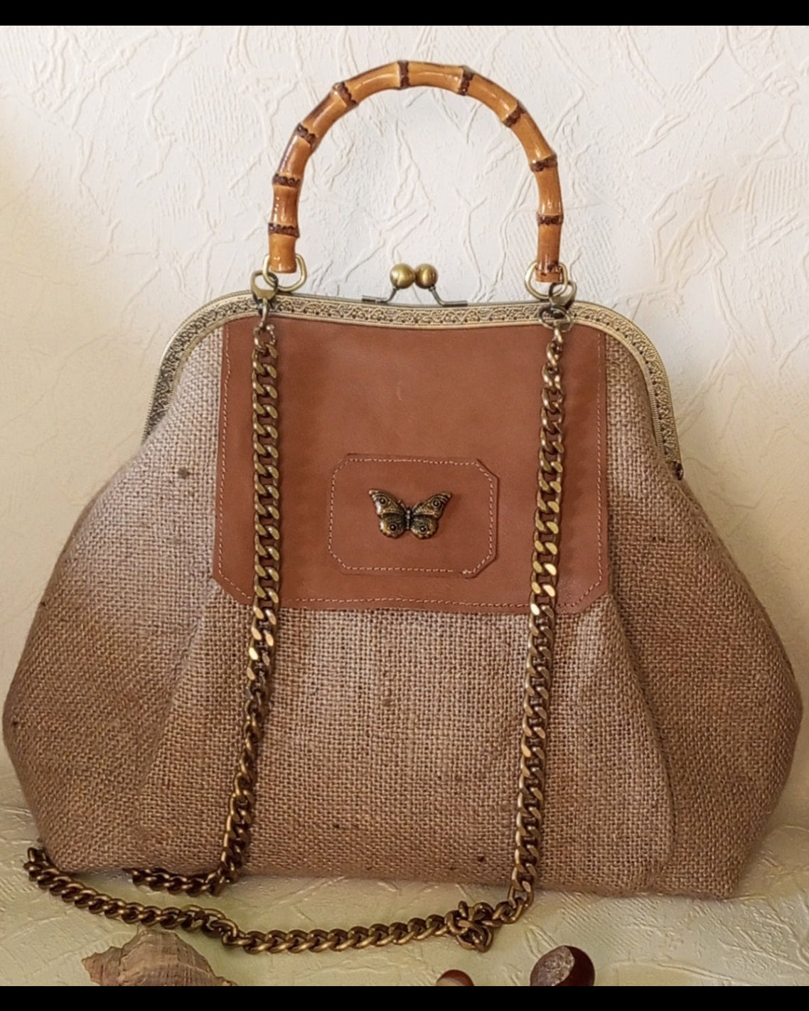 Brown handbag with woven texture, bamboo handle, and butterfly emblem on a textured beige background.