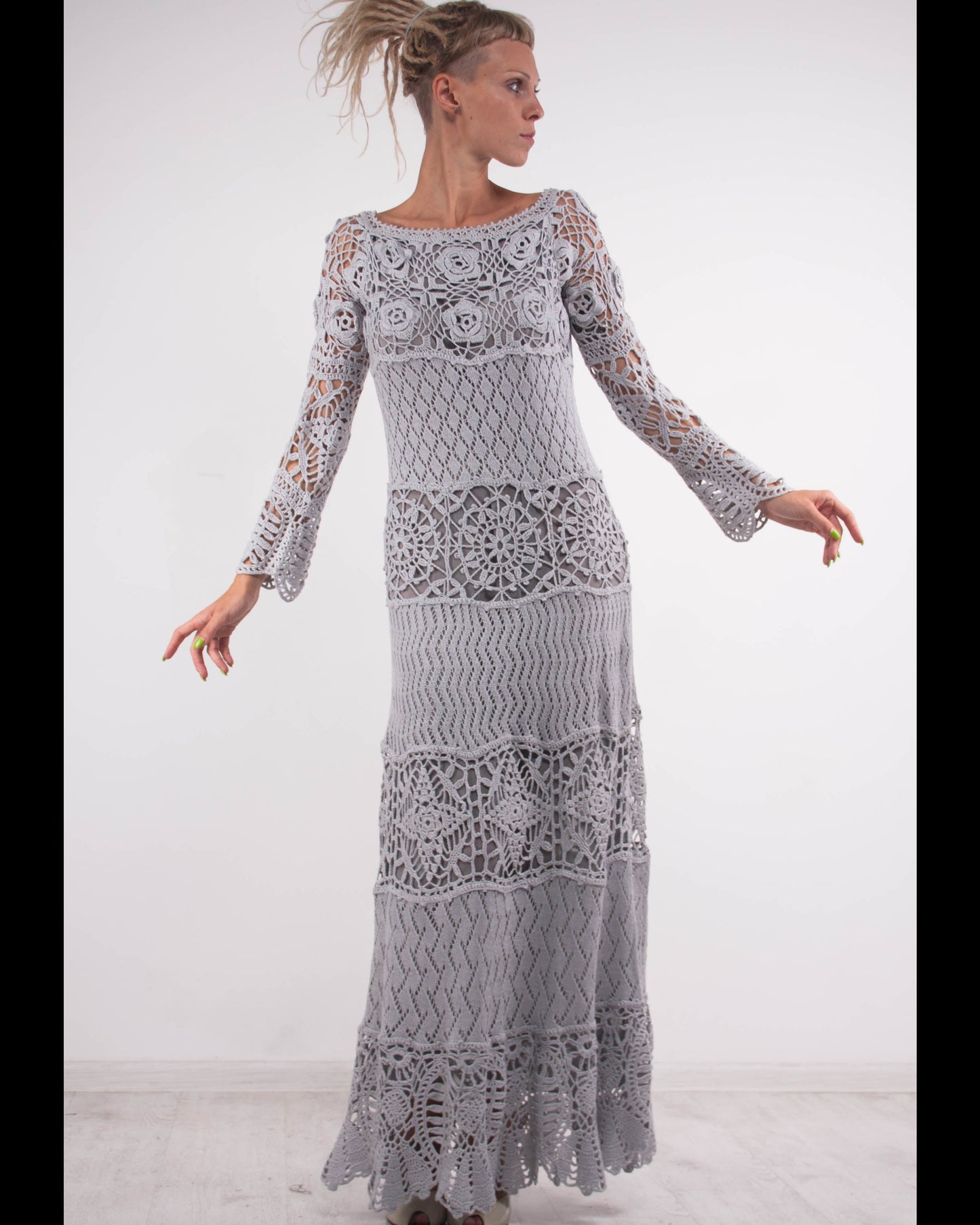 Woman wearing a long, grey, intricately patterned crochet maxi dress against a white background