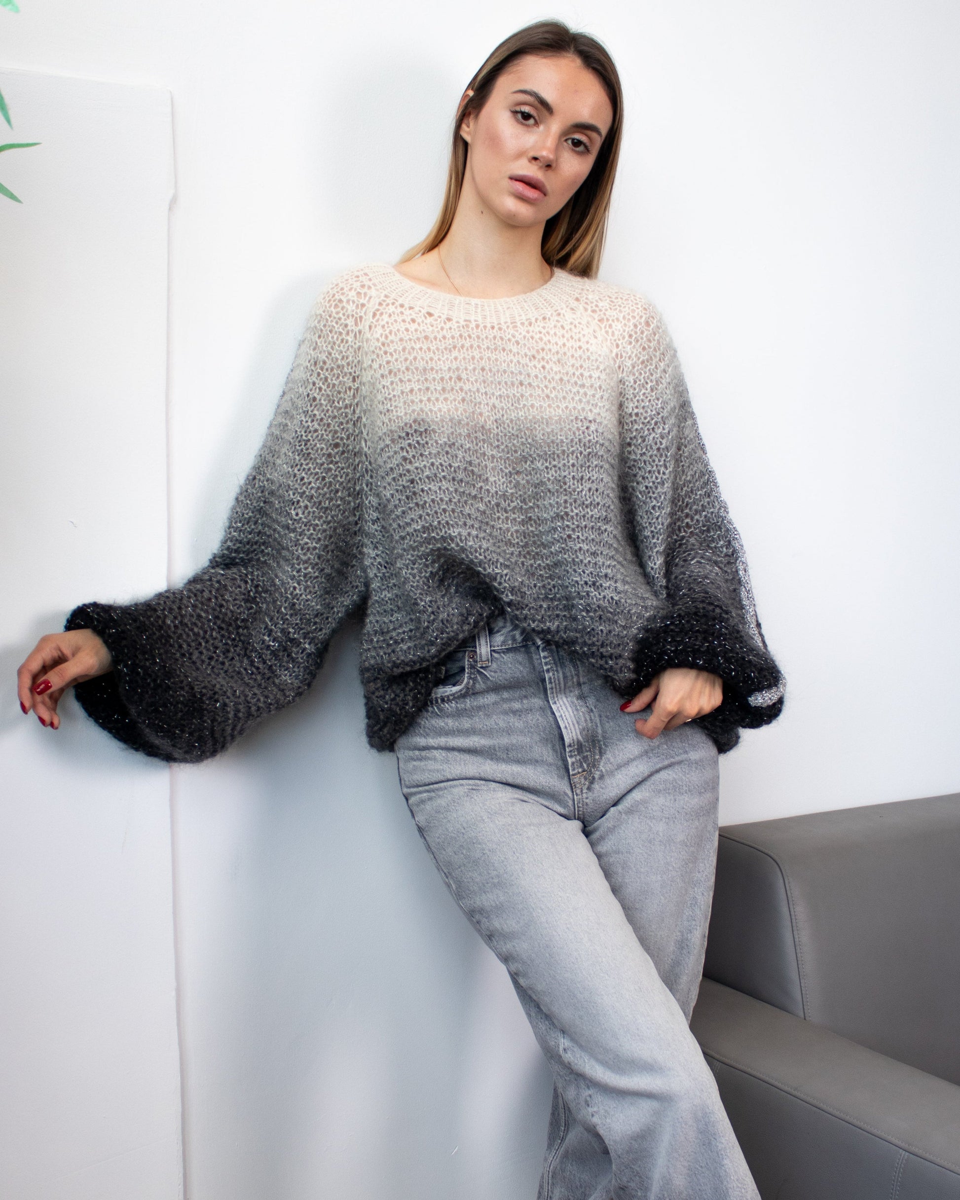 Woman in a gray sweater and jeans leaning against a white wall with a plant.