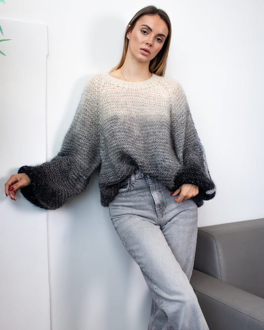 Woman in a gray sweater and jeans leaning against a white wall with a plant.