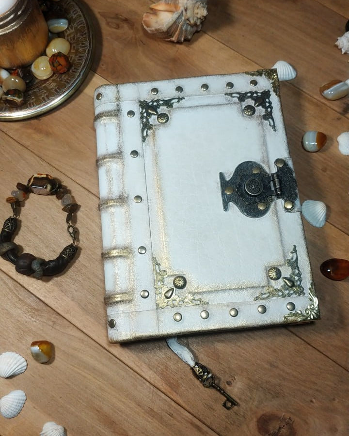 Decorative white aged leather notebook with metal embellishments on a wooden surface with seashells and a bracelet.